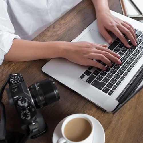 A close-up of hands typing on a laptop placed on a wooden table, with a camera and a cup of coffee also set on the table.