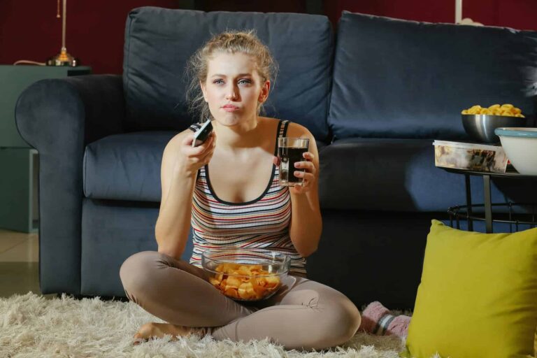 A young woman snacks on ultra-processed foods while watching TV at night, unaware of how to avoid them and make healthier choices.