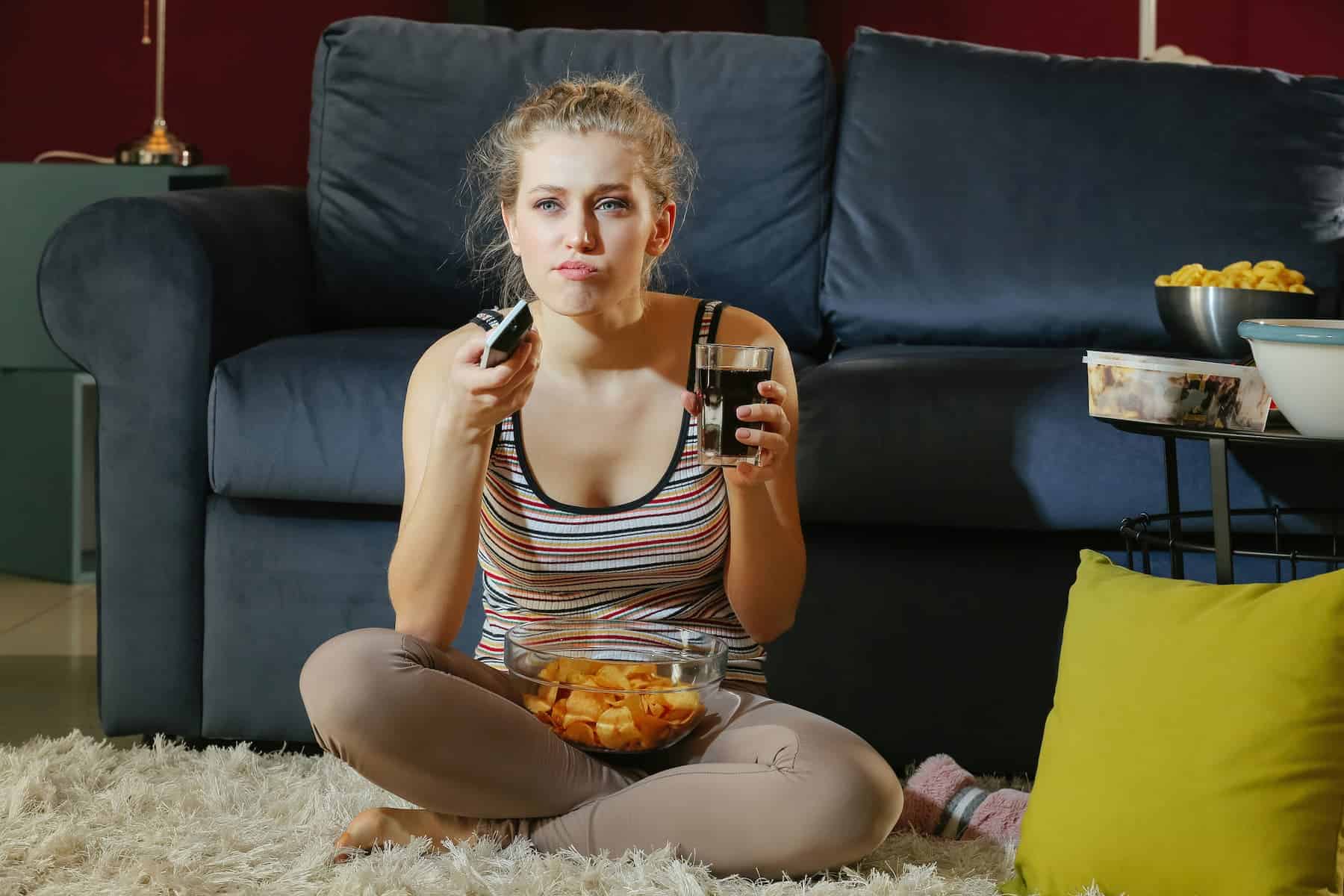 A young woman snacks on ultra-processed foods while watching TV at night, unaware of how to avoid them and make healthier choices.