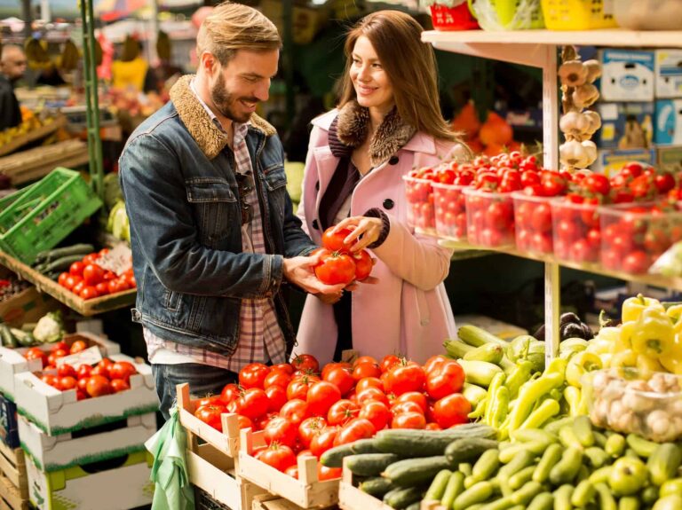A couple is selecting tomatoes at a market produce stand, highlighting fresh items commonly found on a Mediterranean diet food list.