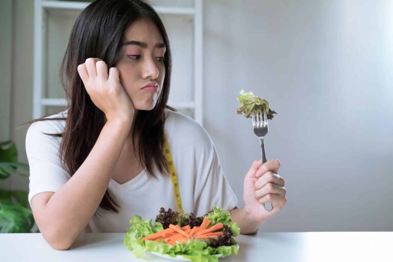 A depressed woman sits alone with a small plate of salad, unaware of the truth about low calorie diets and that 1500 calories is unsustainable for weight loss.