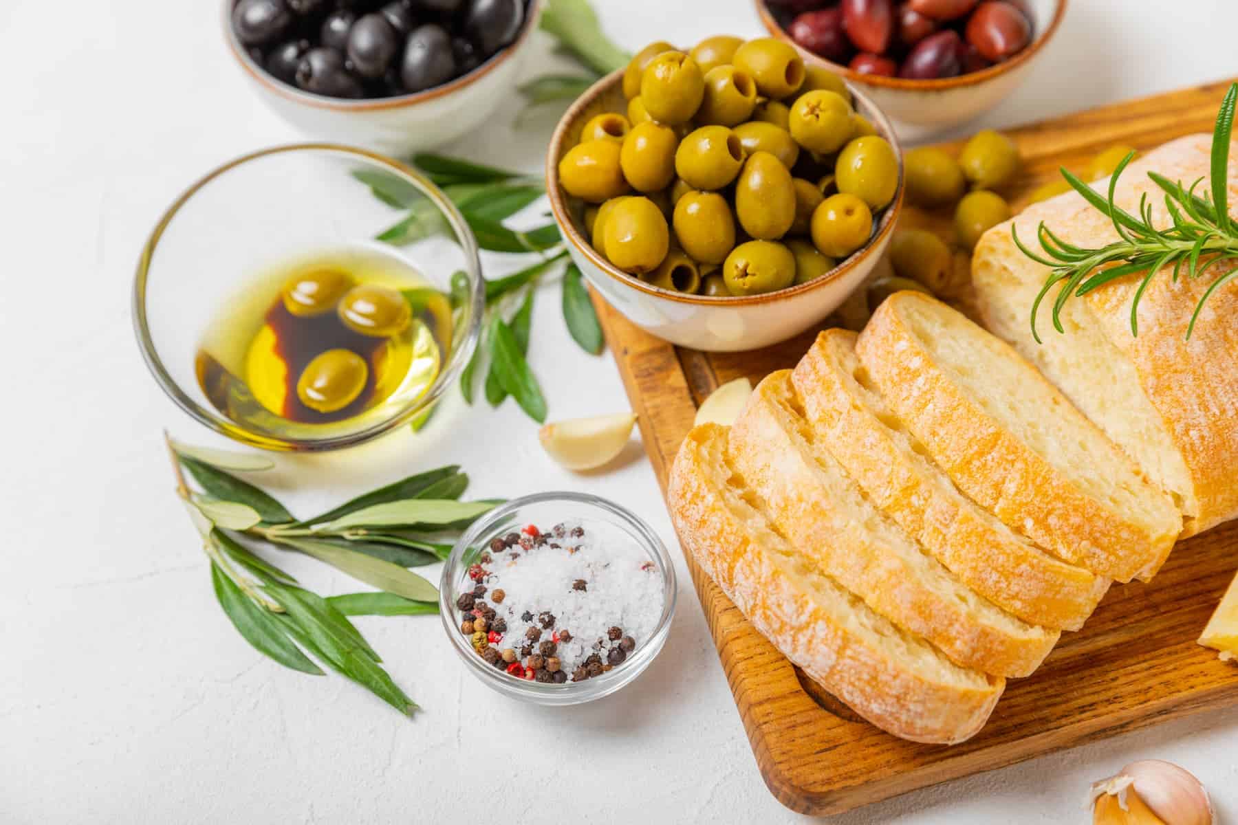 Assorted olives and olive oils in bowls with ciabatta bread on a cutting board, perfect for showcasing types of olive oil.