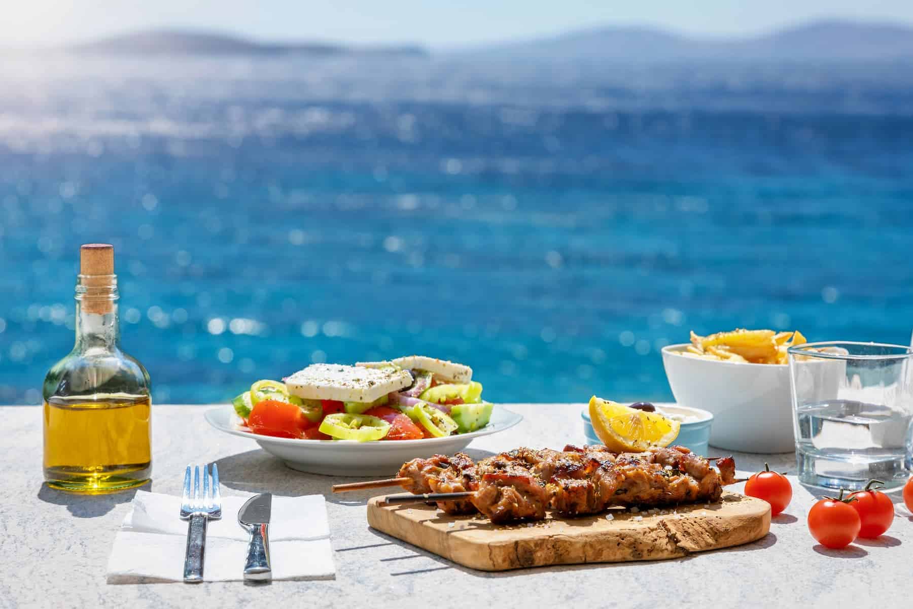 A Greek food scene featuring a fresh salad and a bottle of olive oil, set against the stunning Aegean Sea—highlighting the health benefits of olive oil.