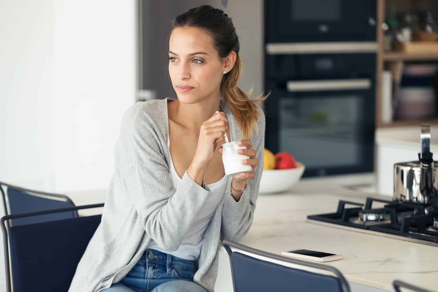 A young woman enjoying yogurt made with her Luvele yogurt maker in the kitchen, with a bowl of fresh fruit behind her, highlighting the importance of probiotics and prebiotics.