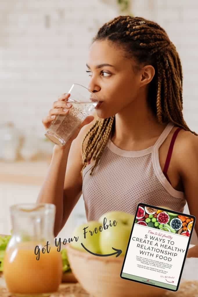 Woman standing in her kitchen and drinking a glass of water. A bowl of apples and fresh juice is in front of her. A small iPad with an image of our free ebook on how to create a healthy food relationship is on the right corner with text that says "get your freebie".