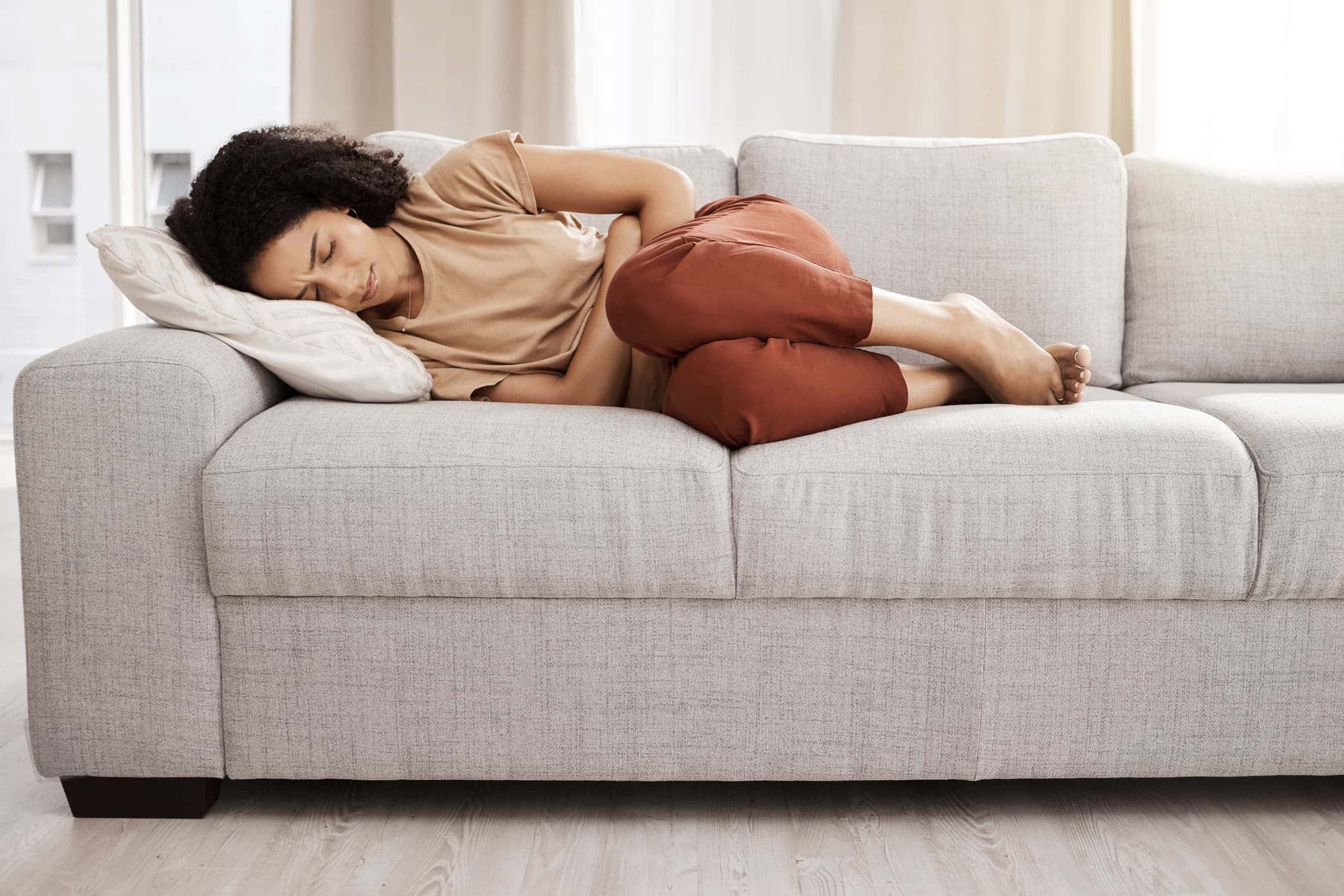 A young woman lying on the sofa at home with stomach pain, realizing the need to improve her gut health.