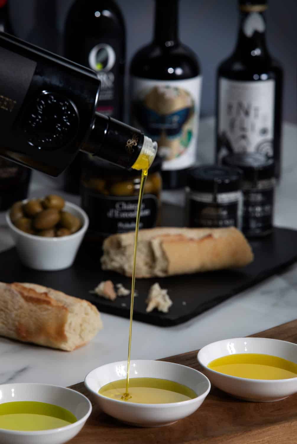 An image of a table with white bread, olives, and a bottle of extra virgin olive oil from Olive Oil Lovers being poured into a small tasting bowl.