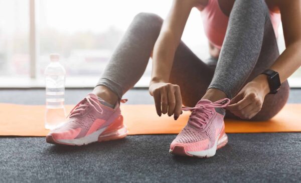 Seated on a yoga mat, a woman in workout clothes ties her pink sneaker, emphasizing preparation for a fitness healthy lifestyle.