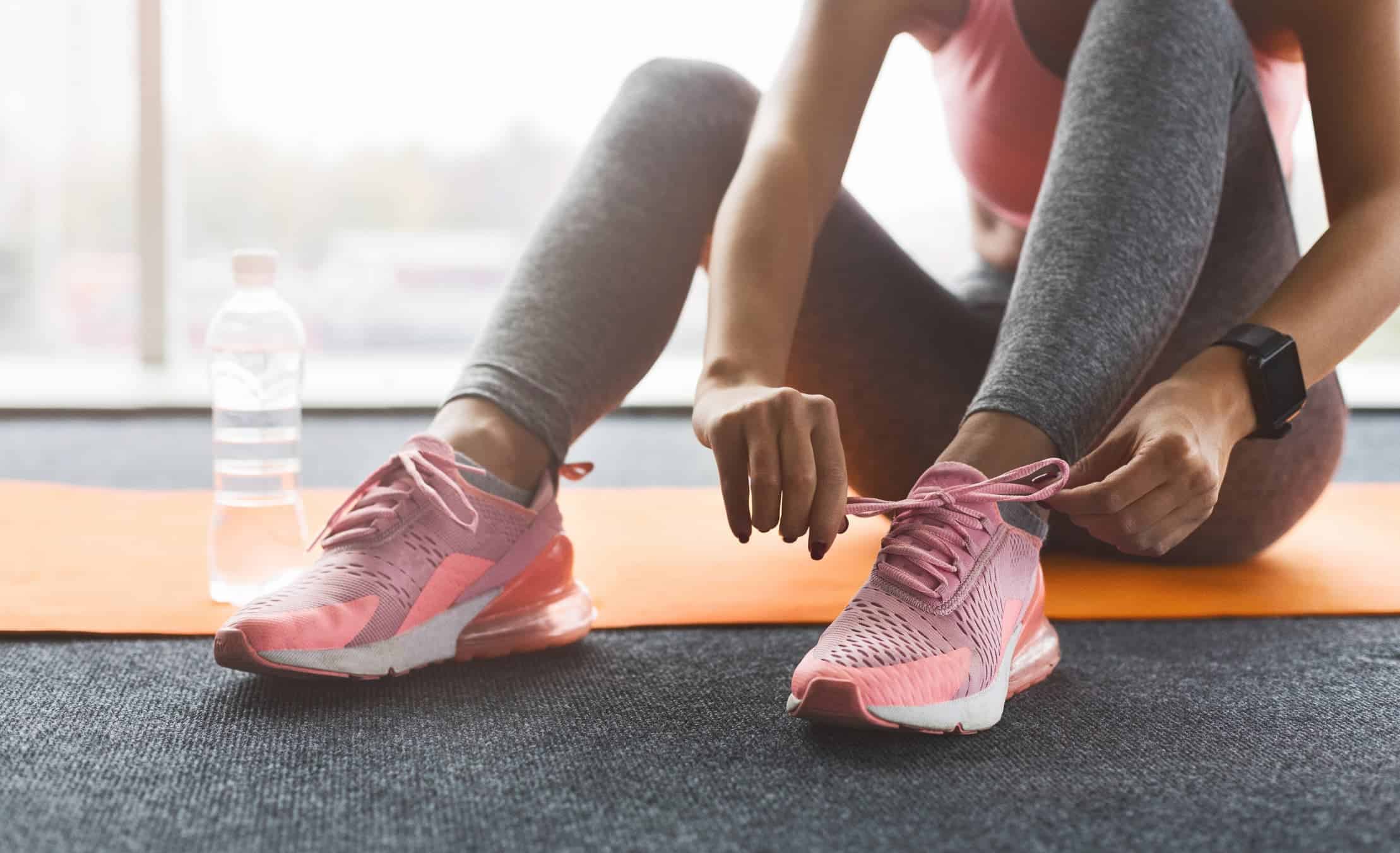 Seated on a yoga mat, a woman in workout clothes ties her pink sneaker, emphasizing preparation for a fitness healthy lifestyle.