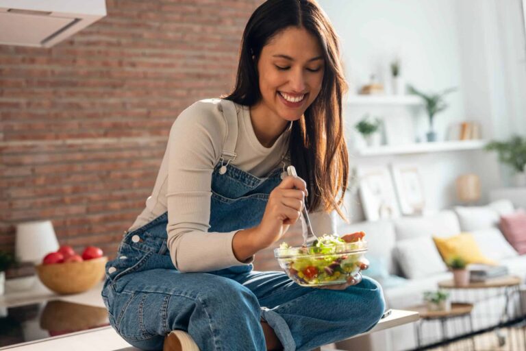 A smiling woman in denim overalls eats a salad from a clear bowl, highlighting nutritious food choices in healthy lifestyle.