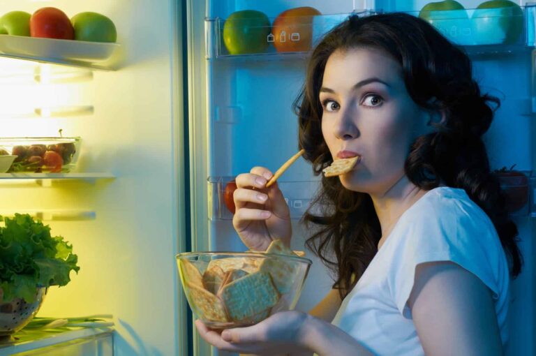 Woman looking guilty as she eats crackers in front of an open fridge at night, wondering why she's always hungry.