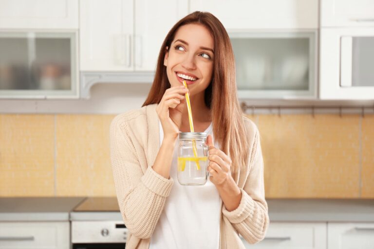 A woman drinks lemon water through a straw in her kitchen, wondering if the benefits of lemon water are real.