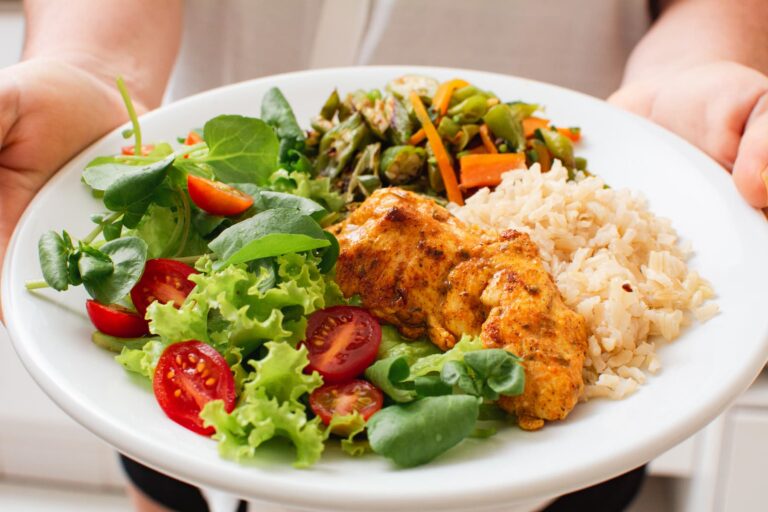 Balanced healthy meal with grilled chicken, rice, vegetables, and salad on a white plate carried by a woman—illustrating the importance of eating enough for sustainable weight loss.