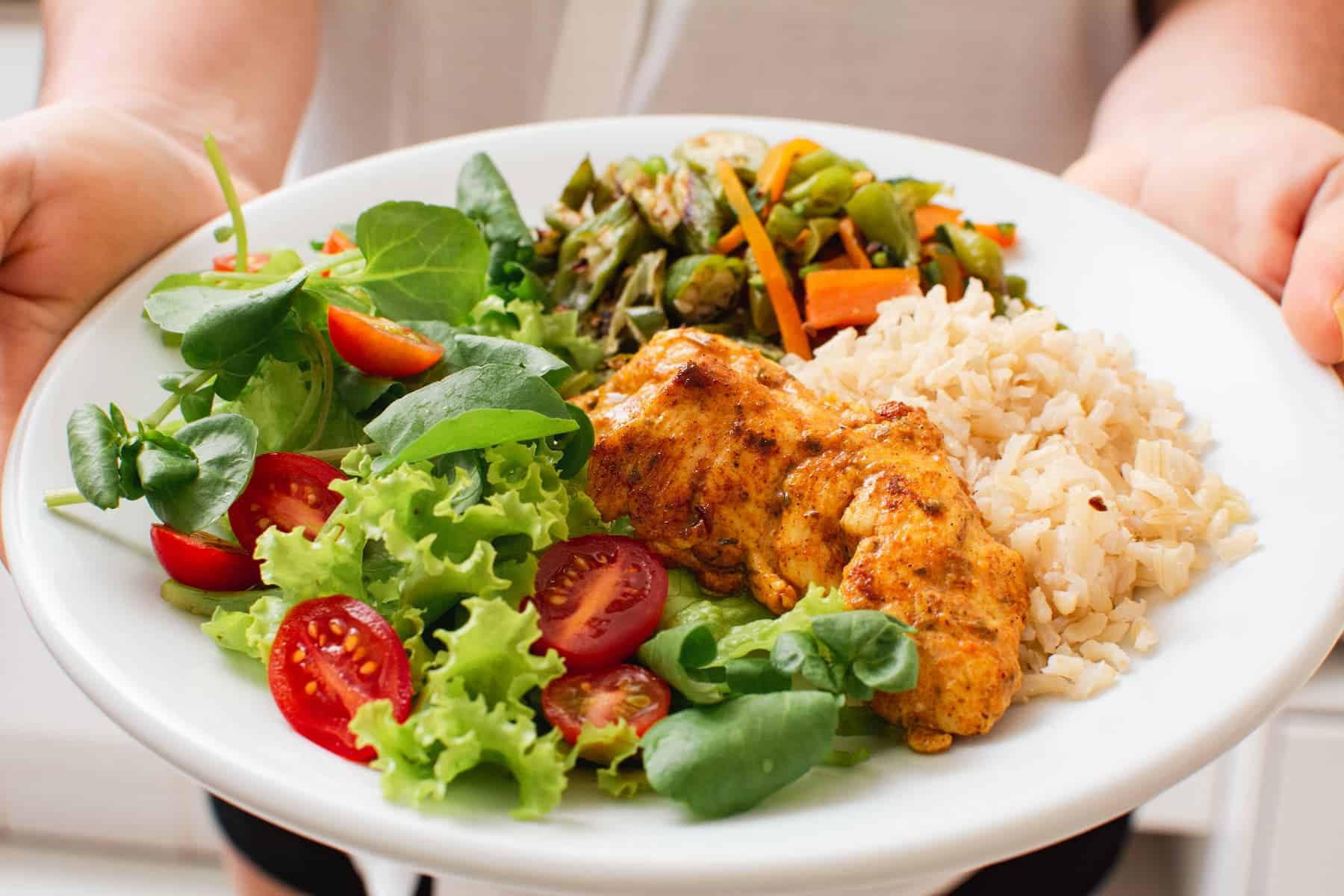 Balanced healthy meal with grilled chicken, rice, vegetables, and salad on a white plate carried by a woman—illustrating the importance of eating enough for sustainable weight loss.