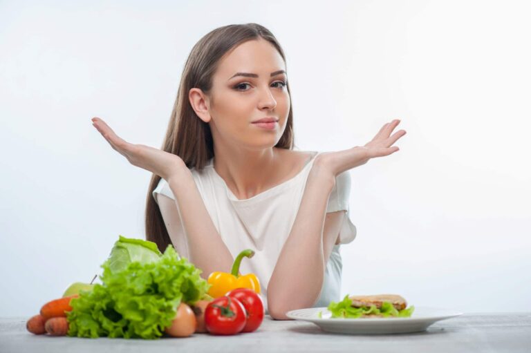 Woman at table with vegetables and sandwich raising hands in question, comparing Mediterranean diet vs. keto choices.