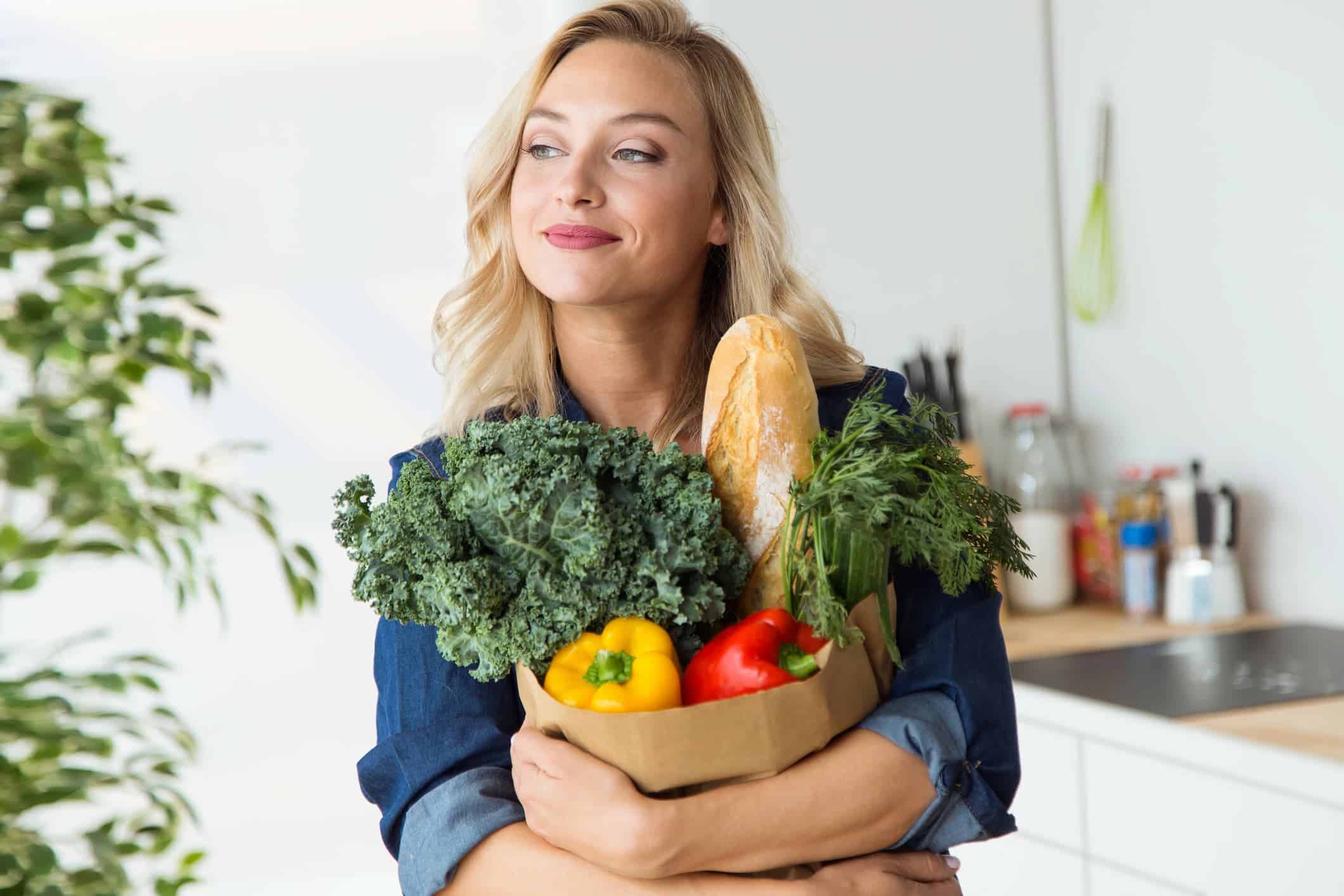 A woman in a blue shirt is holding a grocery bag filled with Mediterranean foods, having embraced the Mediterranean diet for a healthier lifestyle.