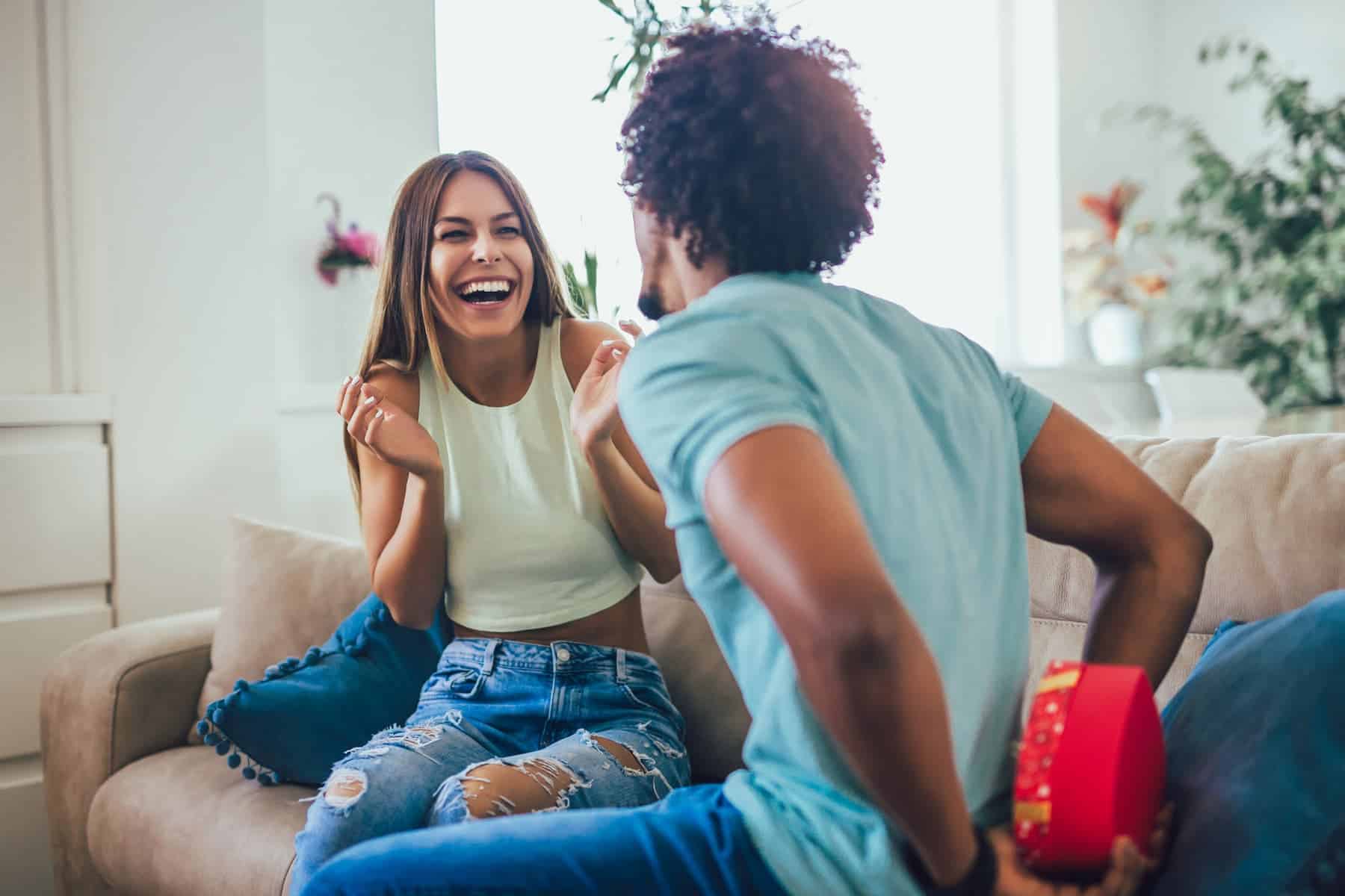 A woman sits on the couch, looking excited as her boyfriend hides a red heart-shaped box behind him, having found a great Valentine’s Day gift idea.