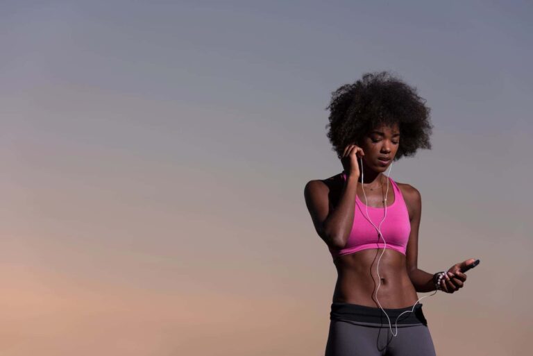 A woman in workout attire listens to music outdoors at sunset while getting ready for her exercise for flat stomach routine.