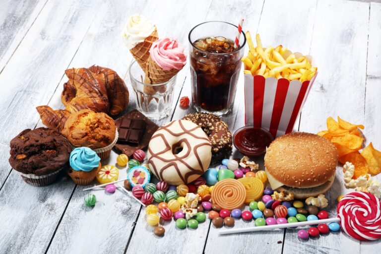 Unhealthy foods that cause inflammation, including burgers, chips, candy, donuts, soda, and ice cream, displayed on a table.