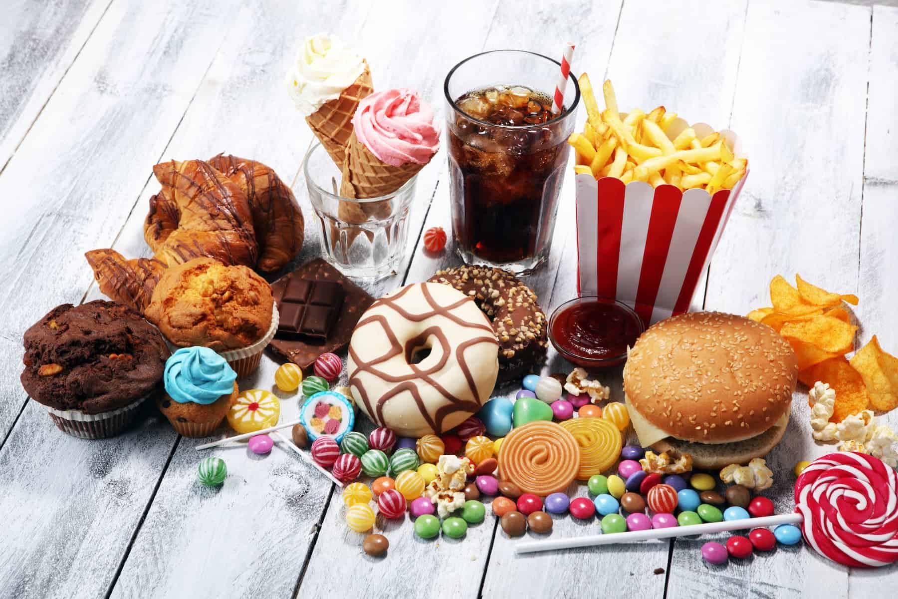 Unhealthy foods that cause inflammation, including burgers, chips, candy, donuts, soda, and ice cream, displayed on a table.