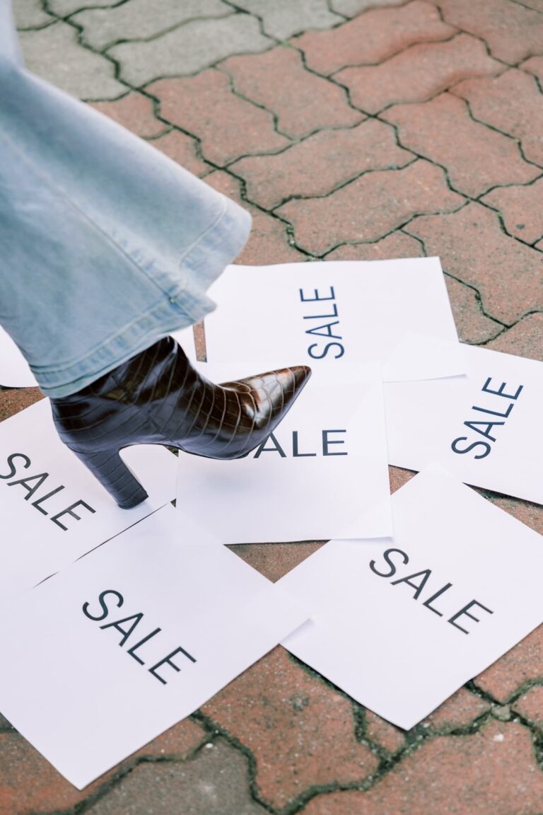 A pointed-toe high-heeled boot steps on papers with the word SALE printed on them highlighting Black Friday deals.