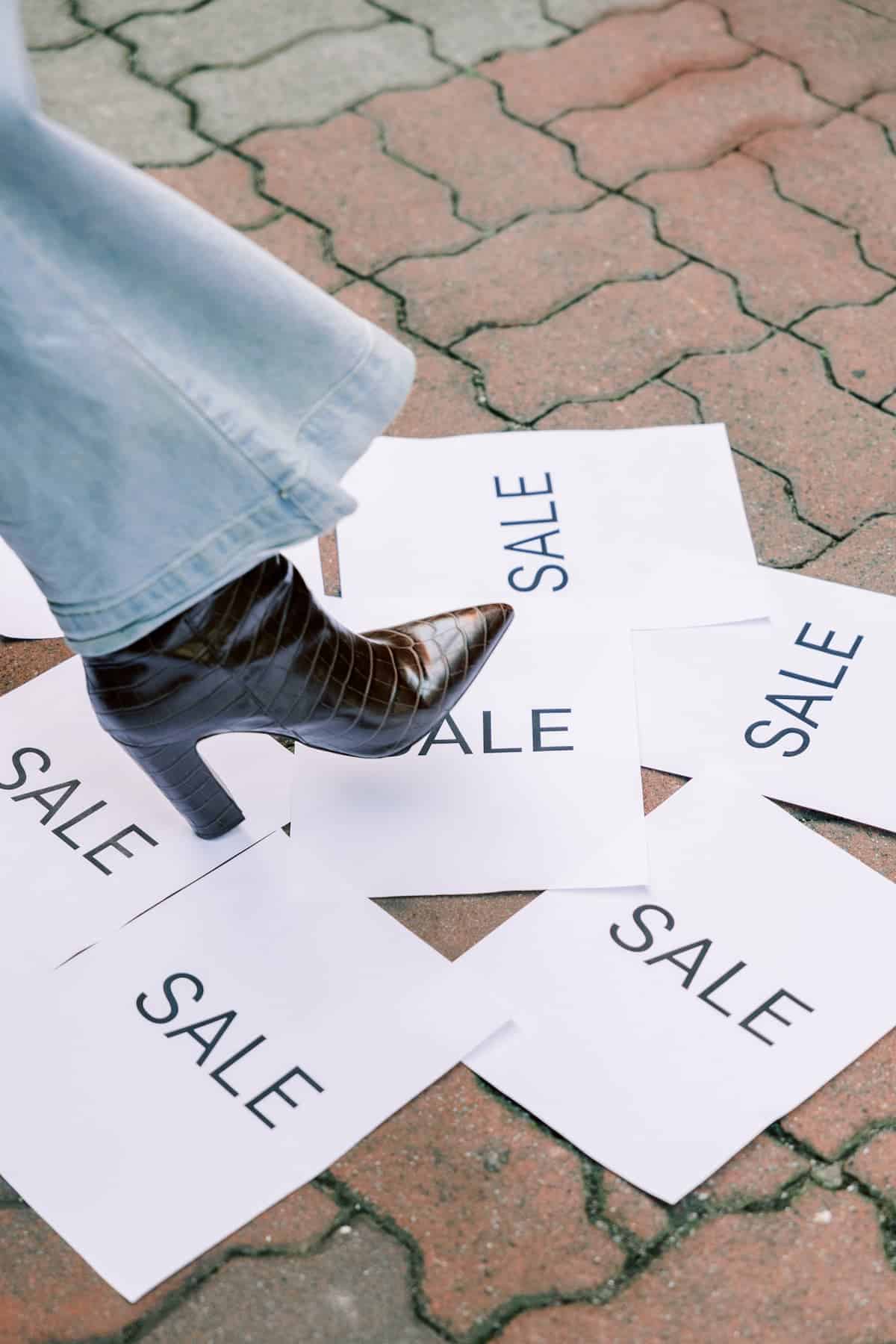 A pointed-toe high-heeled boot steps on papers with the word SALE printed on them highlighting Black Friday deals.