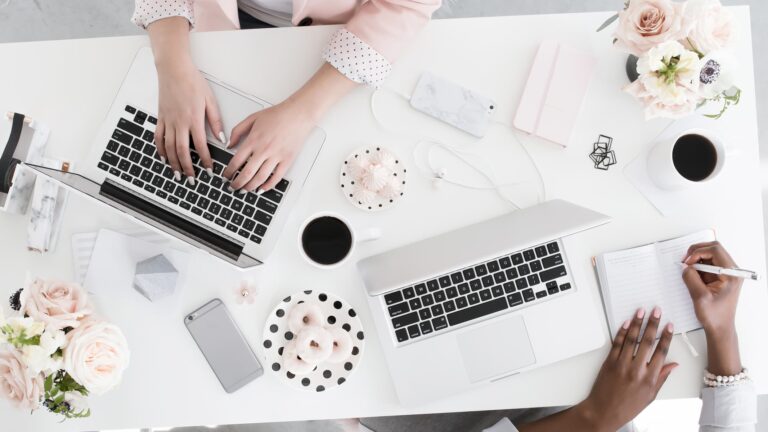 Blogging setup workspace with two women working on laptops at a desk with notebooks, coffee mugs, and office supplies.