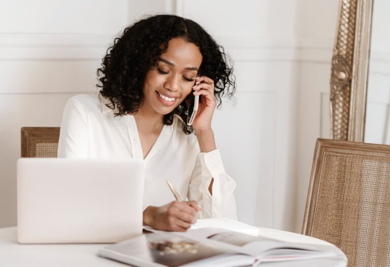 Woman working on a laptop while taking notes and talking on the phone, representing managing a website with premium web hosting.