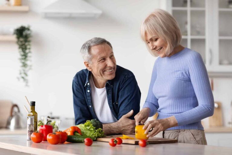 An older couple prepares vegetables together in a kitchen with fresh produce on the counter.