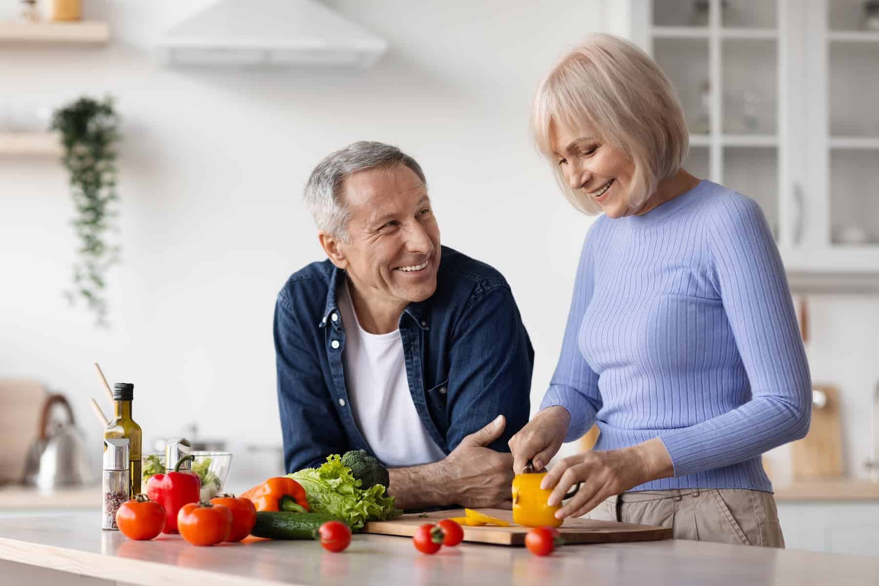 An older couple prepares vegetables together in a kitchen with fresh produce on the counter.