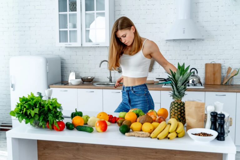 A woman standing in a kitchen in front of a table of fresh fruits and vegetables, illustrating how to get a flat stomach.