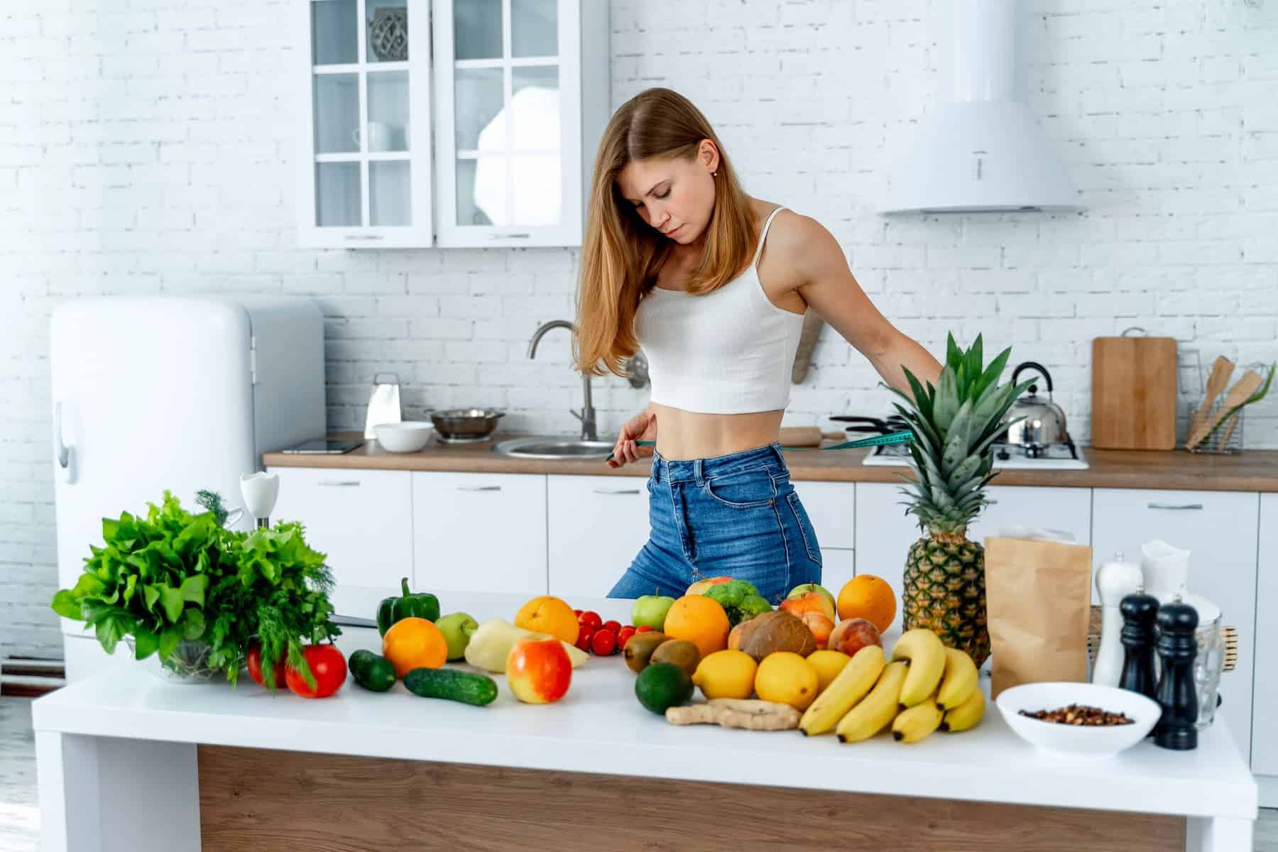 A woman standing in a kitchen in front of a table of fresh fruits and vegetables, illustrating how to get a flat stomach.