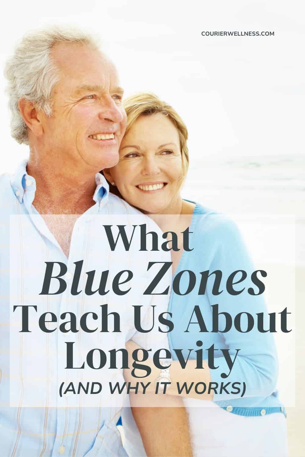 An older couple stands smiling on a beach, with the woman resting her head on the man's shoulder.