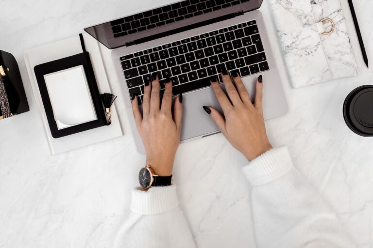 A woman wearing a white sweater and wristwatch types on a laptop at a marble desk with an open notebook and a coffee cup nearby.