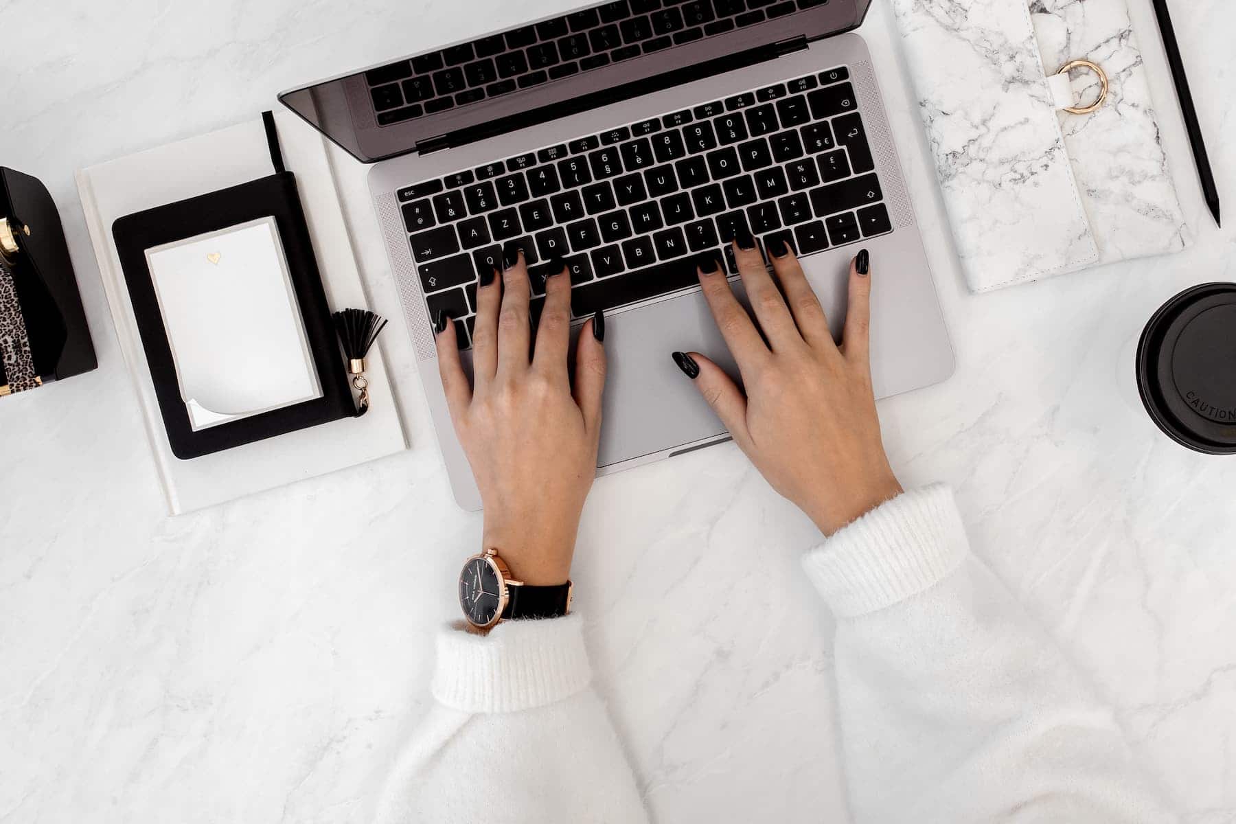 A woman wearing a white sweater and wristwatch types on a laptop at a marble desk with an open notebook and a coffee cup nearby.