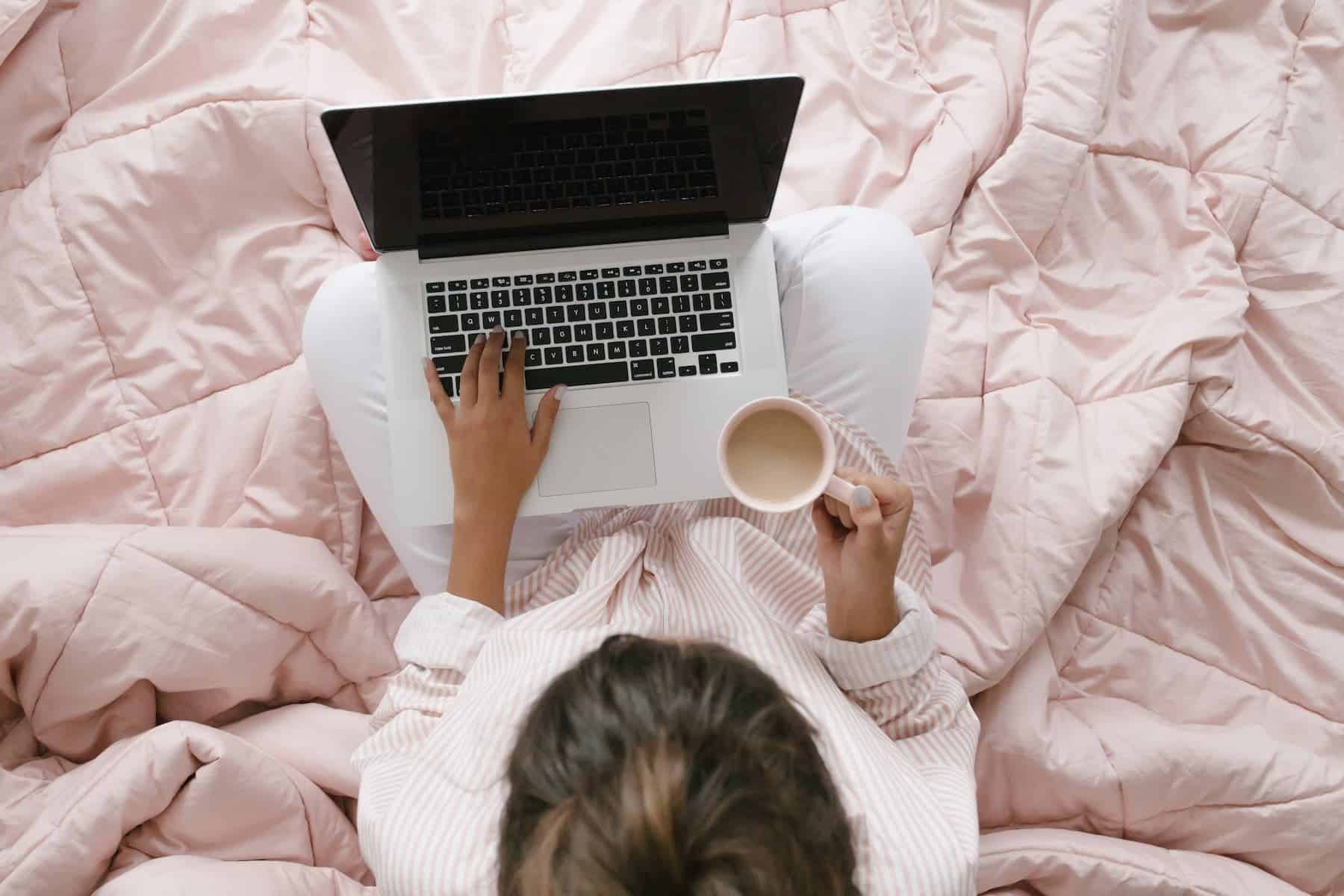 A woman sits on a bed with a pink comforter, typing on a laptop while holding a mug of coffee.